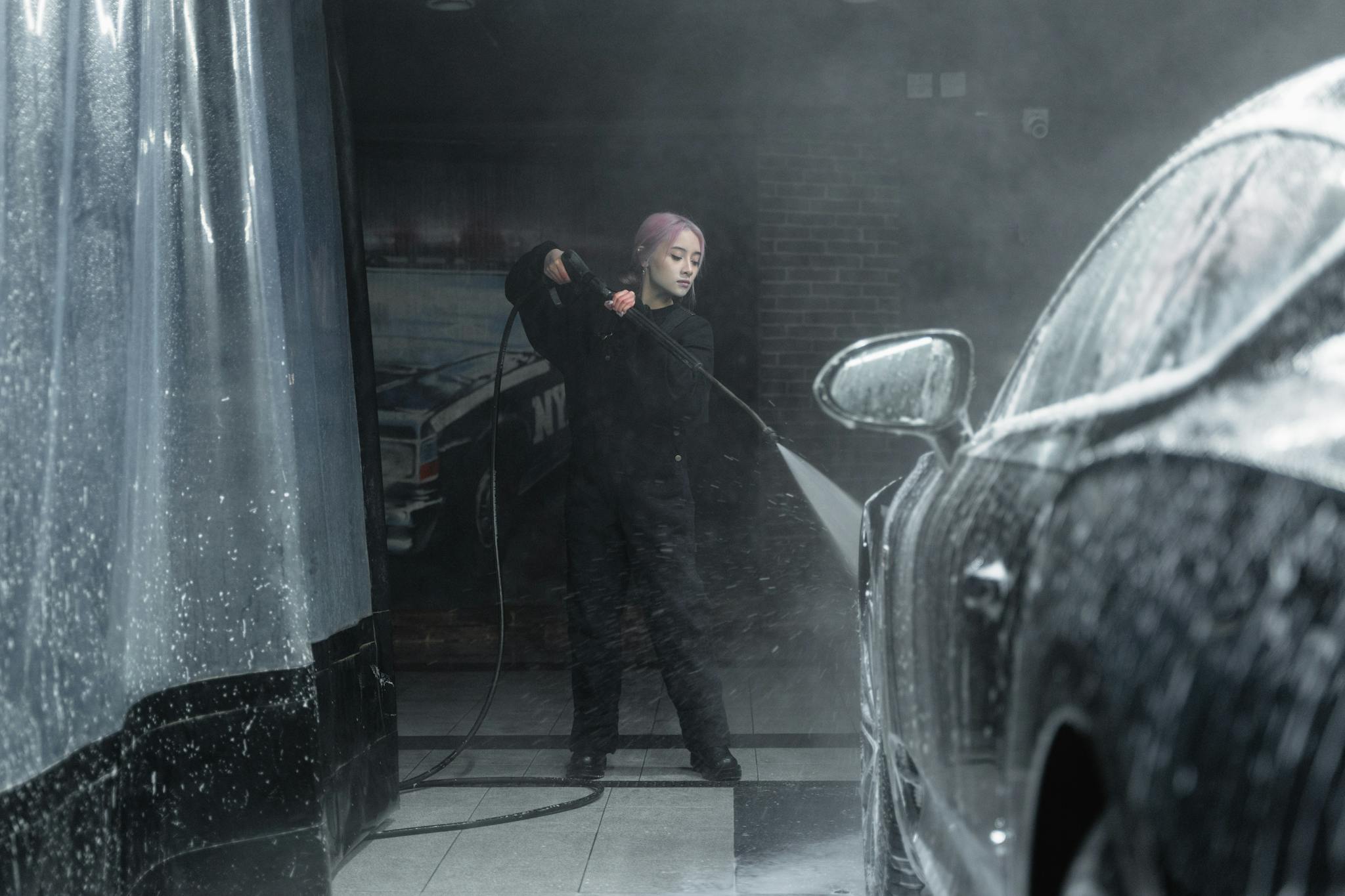 A young woman in a black outfit washing a car with foam and water spray indoors.